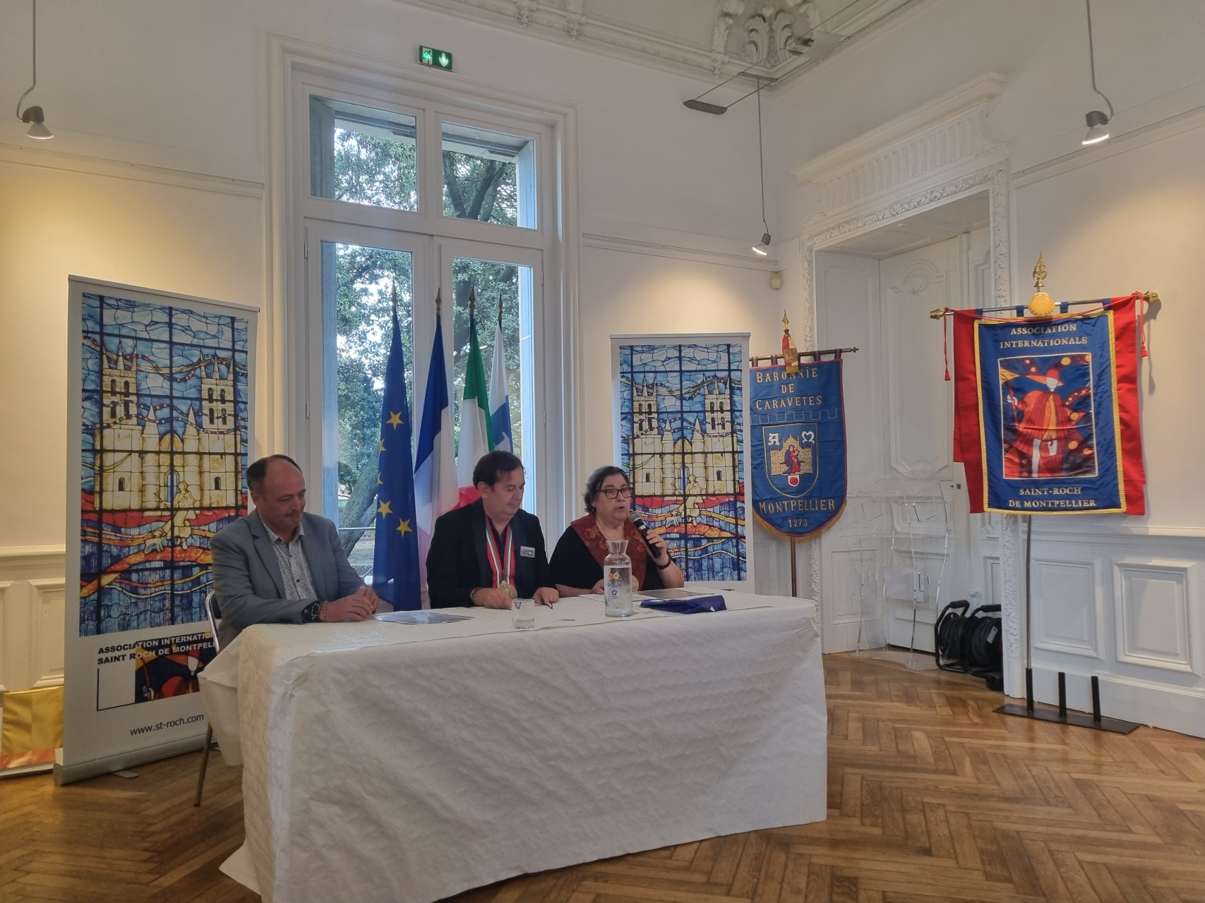 3 personnes assises devant des étendards de Saint Roch de Montpellier, les drapeaux de l'UE, de la France et de l'Italie.