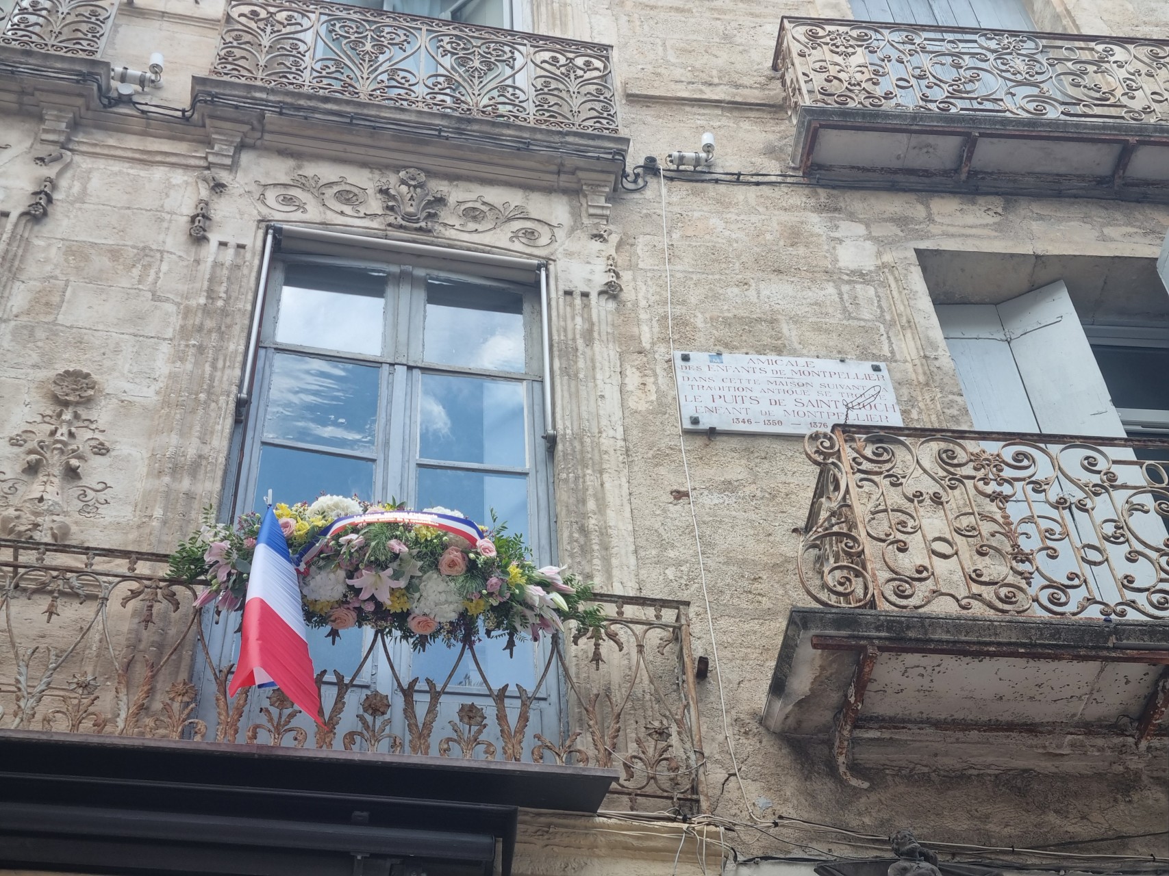 Gerbe de fleurs sur un balcon à côté d'une plaque indiquant le puits de Saint Roch