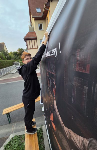 Leo debout sur un banc en tenant à bout de bras la bâche représentant Florian Daguet et son échoppe.